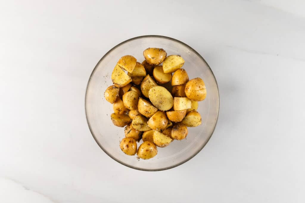 A bowl of seasoned, roasted potato wedges on a light-colored marble surface. The potatoes are cut into small, uniform pieces with herbs and spices visible. The bowl is clear, offering an overhead view of the contents.