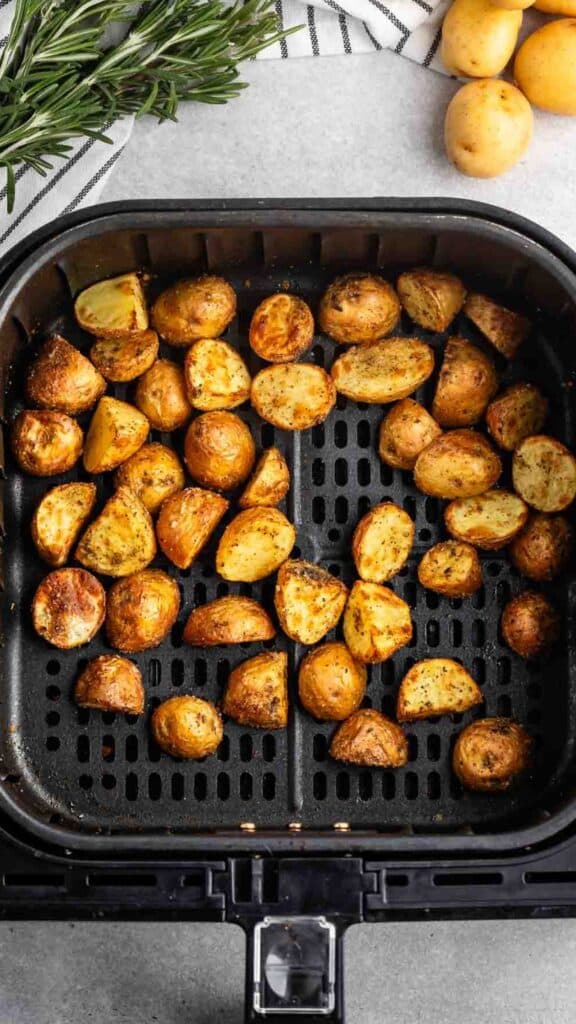 Golden roasted potato halves seasoned with spices in an air fryer basket, surrounded by fresh rosemary, a striped towel, and whole potatoes on a gray surface.