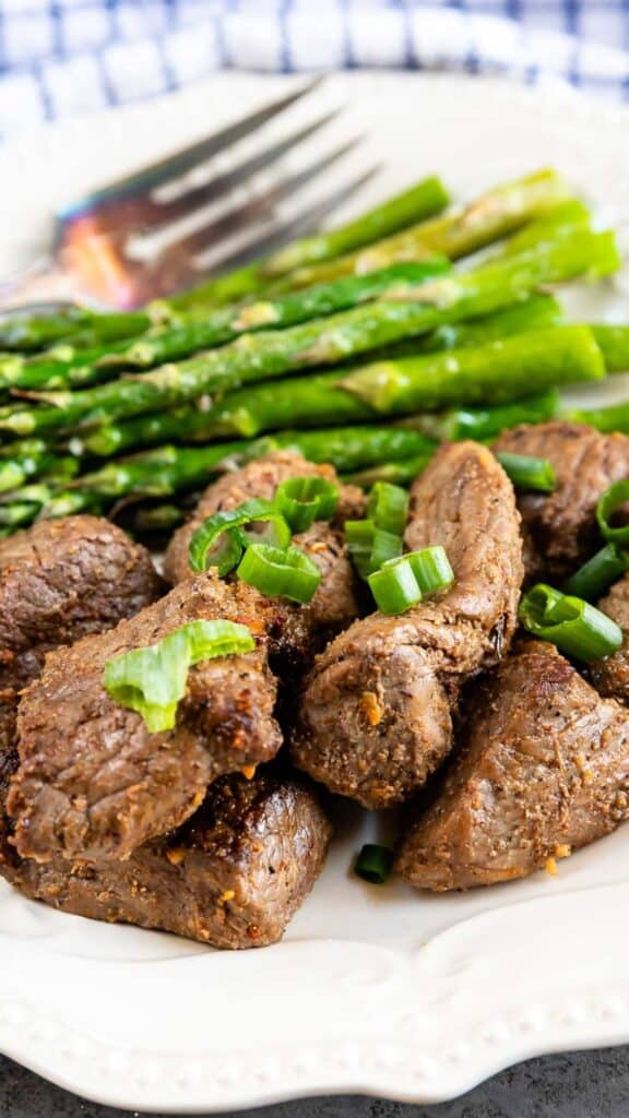 A white plate with seasoned steak bites garnished with chopped green onions, served alongside lightly cooked asparagus spears. A fork is placed in the background on a blue checkered cloth.