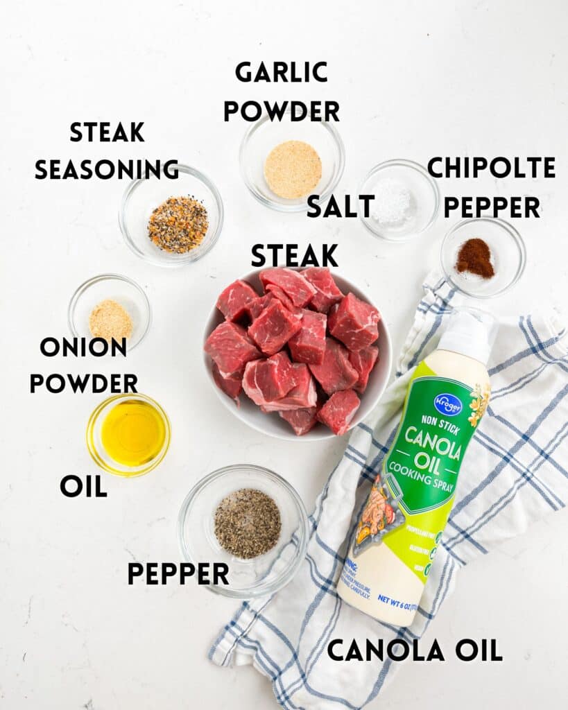 A collection of ingredients is arranged on a white surface, including steak cubes, bowls of garlic powder, salt, chipotle pepper, onion powder, steak seasoning, oil, pepper, and a can of canola oil cooking spray. A striped towel lies nearby.