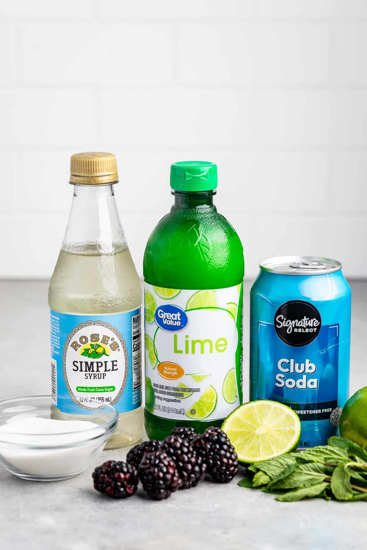 Assorted cocktail ingredients including a bottle of simple syrup, lime juice, and a can of club soda, displayed with fresh blackberries, lime halves, and mint leaves on a gray countertop.