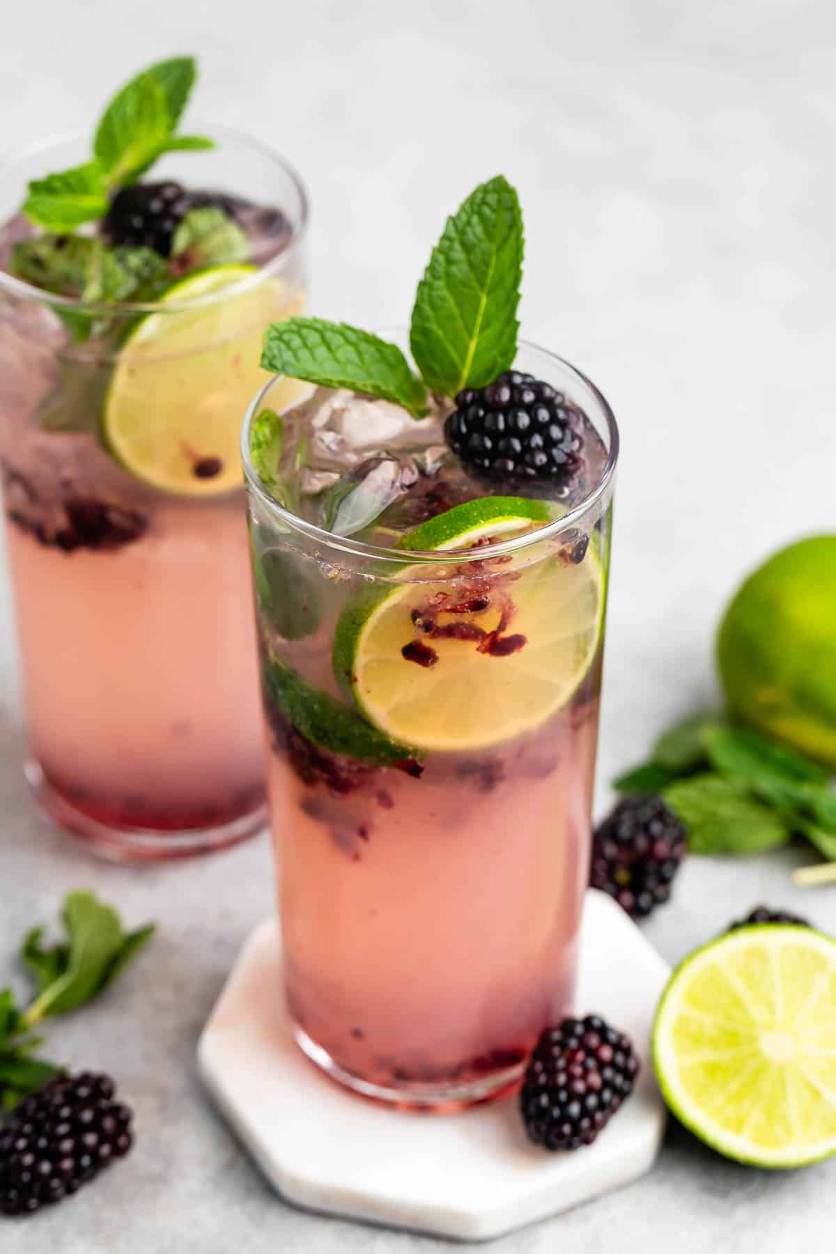 Two refreshing glasses of blackberry mojito with lime slices, mint leaves, and ice cubes. The drinks are garnished with blackberries and mint, surrounded by fresh ingredients on a light surface.