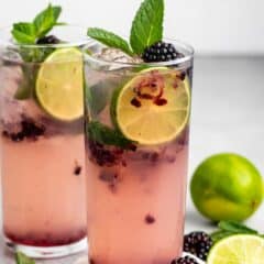 Two tall glasses of a refreshing blackberry lime drink with ice, garnished with mint leaves and lime slices. Fresh blackberries and a halved lime are scattered nearby. The drink has a pastel pink hue, suggesting a fruity, cool beverage.