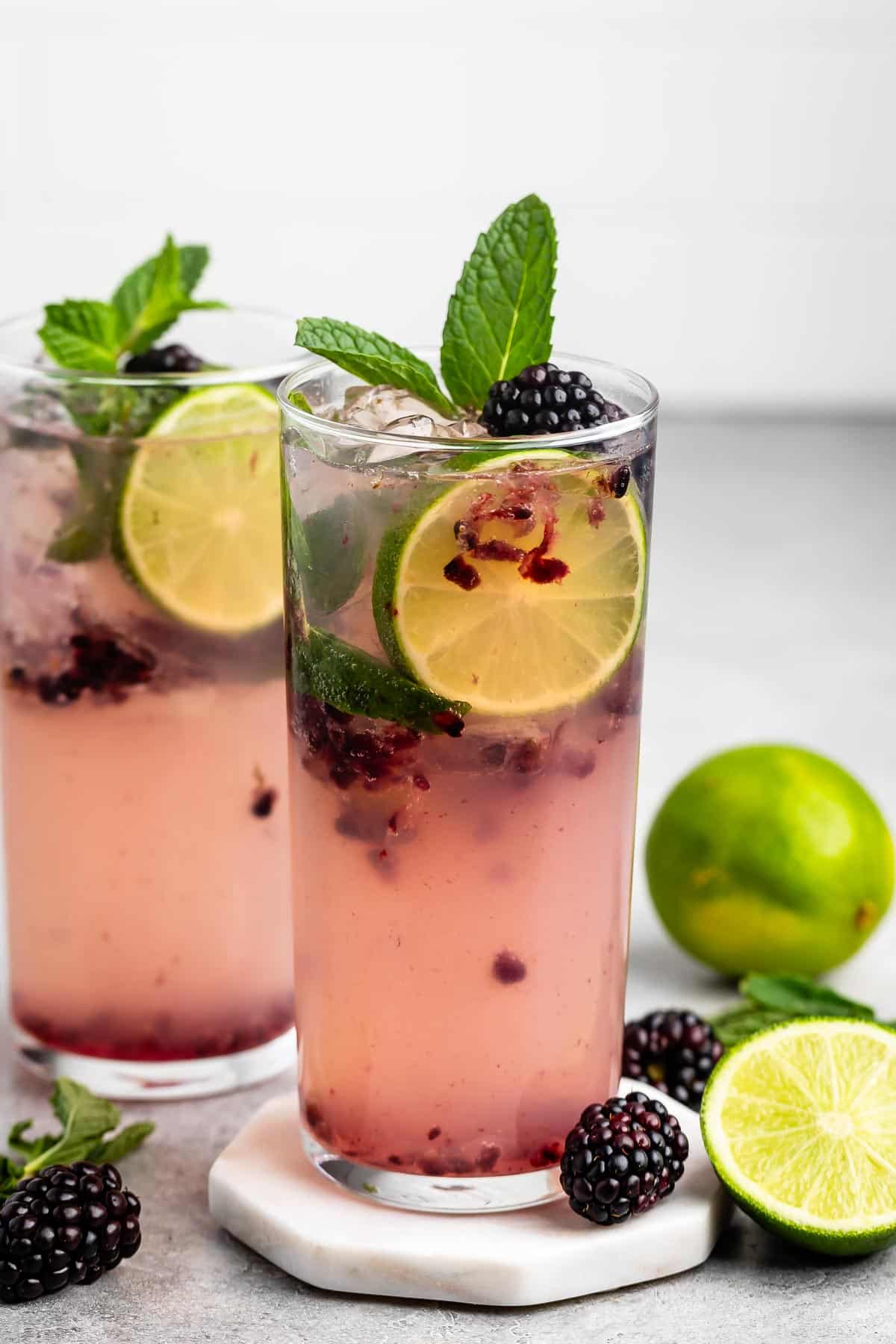 Two tall glasses of a refreshing blackberry lime drink with ice, garnished with mint leaves and lime slices. Fresh blackberries and a halved lime are scattered nearby. The drink has a pastel pink hue, suggesting a fruity, cool beverage.