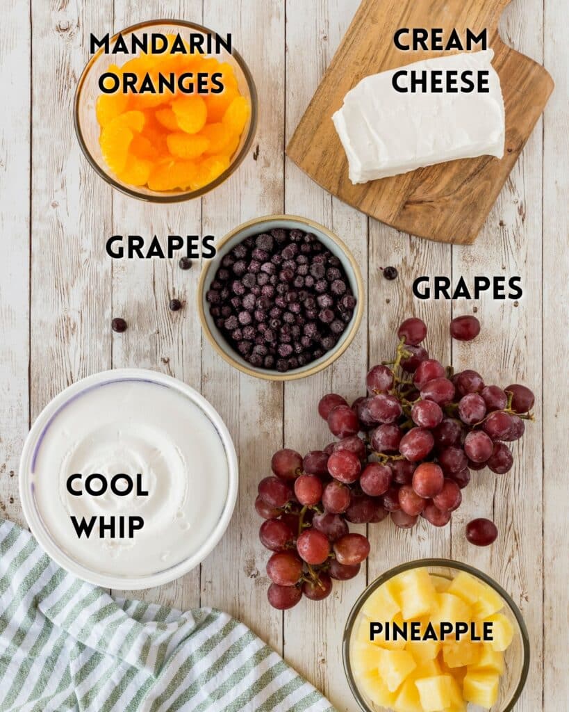 Overhead view of ingredients for a fruit salad on a wooden table: mandarin oranges, cream cheese on a cutting board, black grapes in a bowl, red grapes, a bowl of Cool Whip, and pineapple chunks. A green and white striped cloth is at the bottom left.