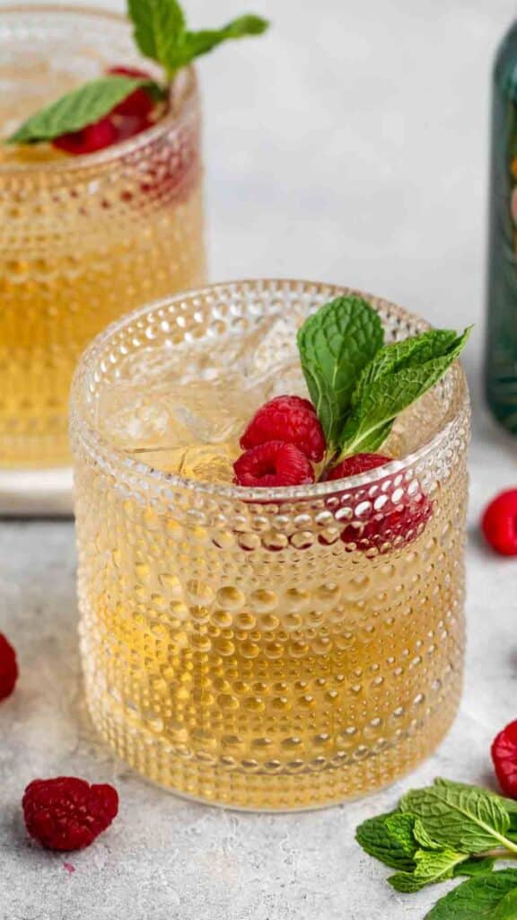 Close-up of a textured glass filled with a light amber beverage, garnished with fresh mint leaves and raspberries. Another similar drink is slightly blurred in the background. Raspberries and mint leaves are scattered on the surface around the glass.