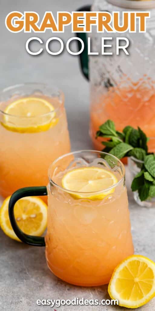 Two glass mugs of grapefruit cooler garnished with lemon slices sit on a table. A jug of the cooler is in the background next to fresh mint leaves. A lemon slice is placed next to the mugs.