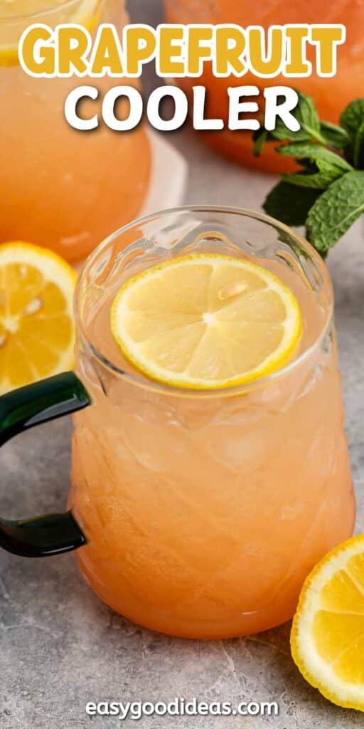 A refreshing grapefruit cooler in a textured glass mug with a lemon slice garnish. Surrounded by additional lemon slices, a full glass pitcher of the drink, and fresh mint leaves on a gray surface. Grapefruit Cooler text on top.