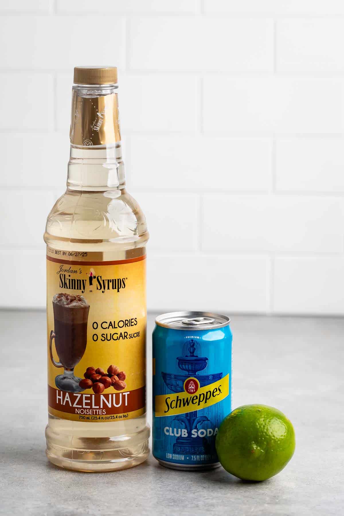 A bottle of Jordans Skinny Syrups Hazelnut flavor, a can of Schweppes club soda, and a lime are arranged on a gray surface. The background features a white tiled wall.