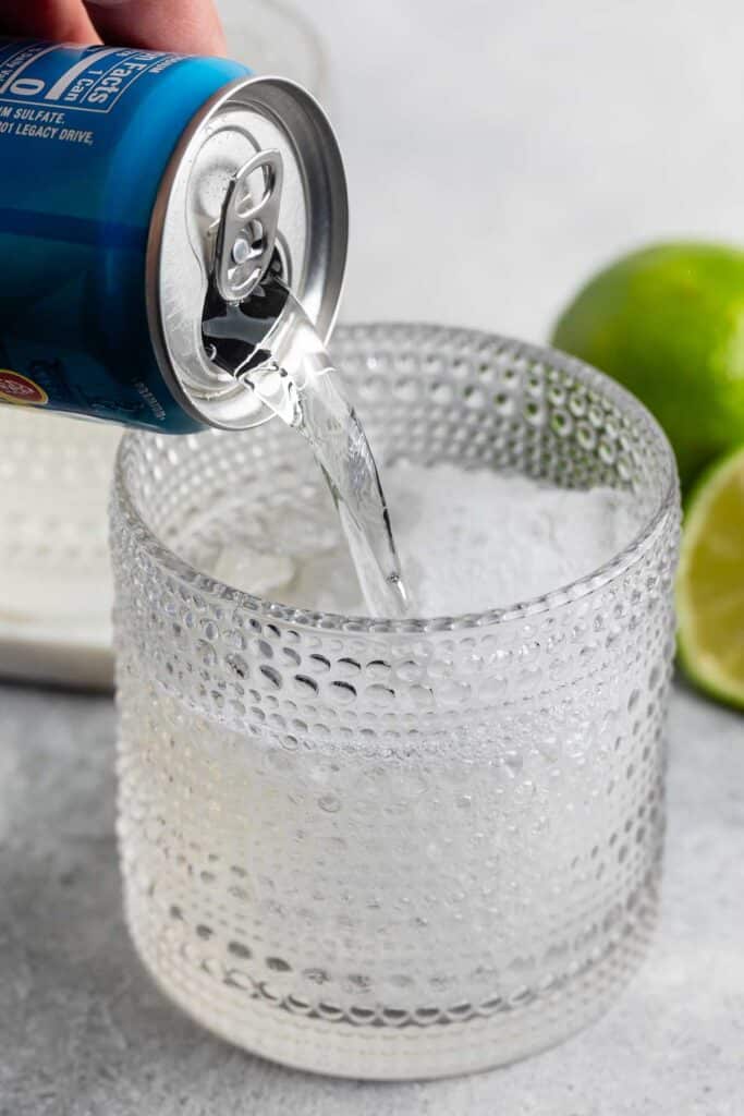 A hand pours a clear soda from a blue can into a textured glass filled with ice. Two lime halves are visible in the background on a light-colored surface.