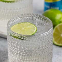 A textured glass filled with a clear, fizzy drink is garnished with a lime slice. In the background, there are whole and half-cut limes, and a blue bottle is partially visible.