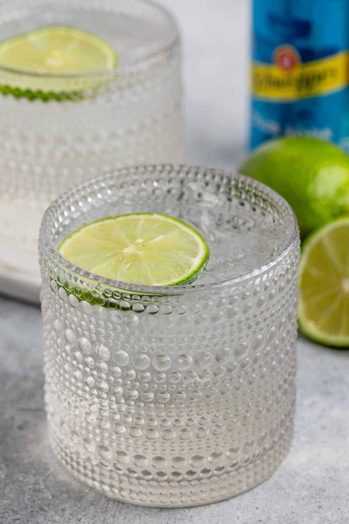 A textured glass filled with a clear, fizzy drink is garnished with a lime slice. In the background, there are whole and half-cut limes, and a blue bottle is partially visible.