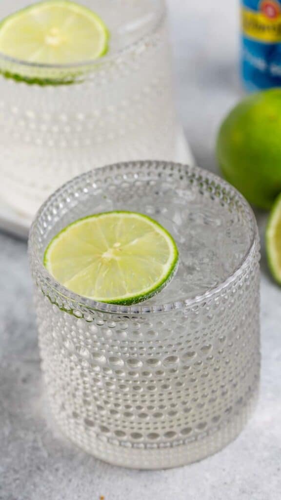 A textured glass filled with a clear beverage and garnished with a lime slice. Another similar glass and whole limes are in the background on a light surface.