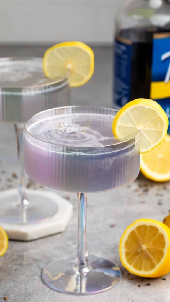 Two iridescent cocktail glasses on a table, each filled with a lavender-hued drink, garnished with a lemon slice. Fresh lemon halves and a bottle are visible in the background.