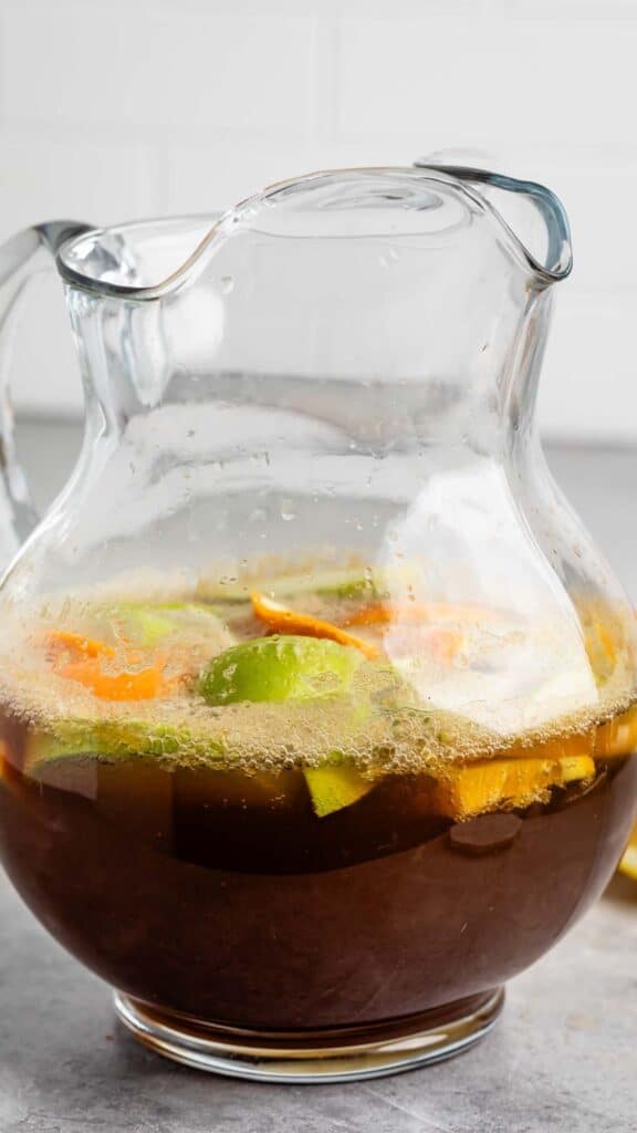 A glass pitcher filled with a dark beverage containing slices of lime, orange, and other fruits. The liquid is frothy at the top, and the pitcher is placed on a gray surface.