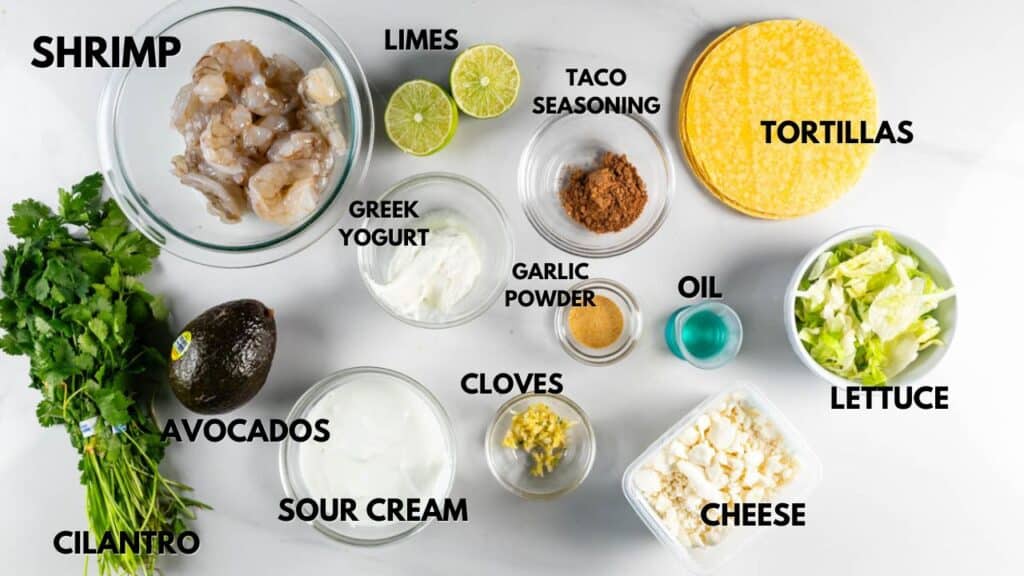 Ingredients for shrimp tacos are spread out: raw shrimp, cilantro, avocados, limes, Greek yogurt, sour cream, garlic powder, taco seasoning, oil, cloves, tortillas, lettuce, and cheese. Each item is labeled.