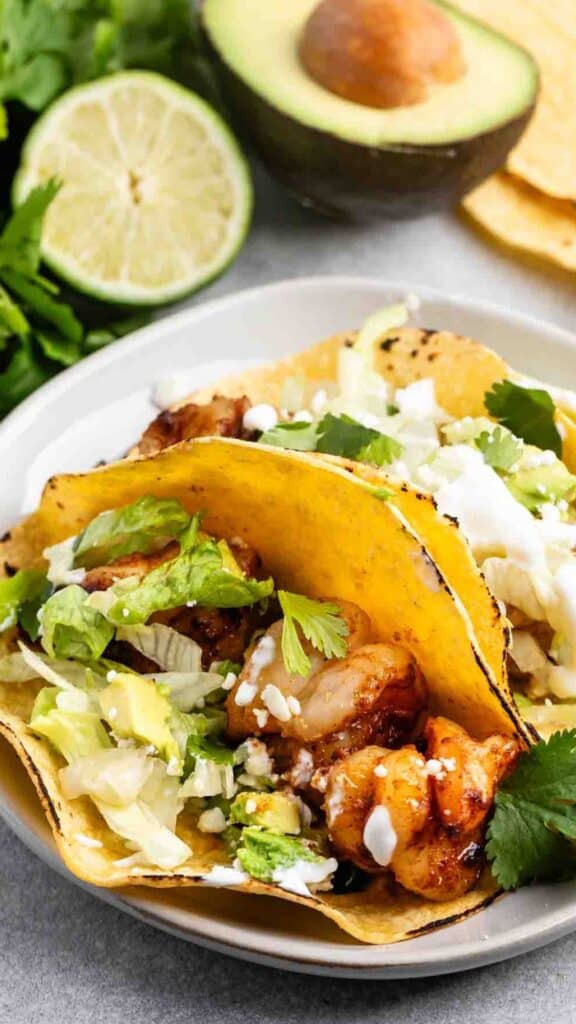 Close-up of two chicken tacos on a plate, filled with grilled chicken, lettuce, and cilantro. A lime half and avocado are in the background, with corn tortillas visible next to the tacos. The scene is bright and appetizing.