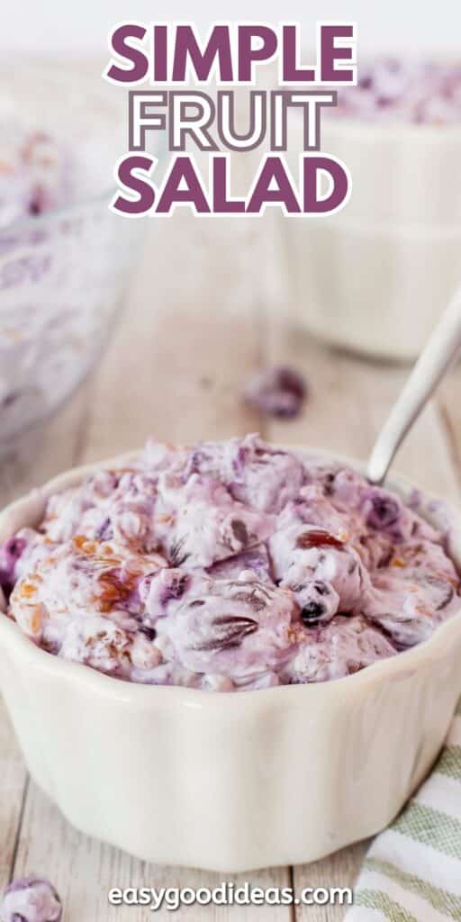 A bowl of creamy fruit salad with blueberries and other fruits, topped with a purple sauce. A spoon is in the bowl. The text Simple Fruit Salad is on top, and a website URL is at the bottom. The background shows another bowl of the salad.