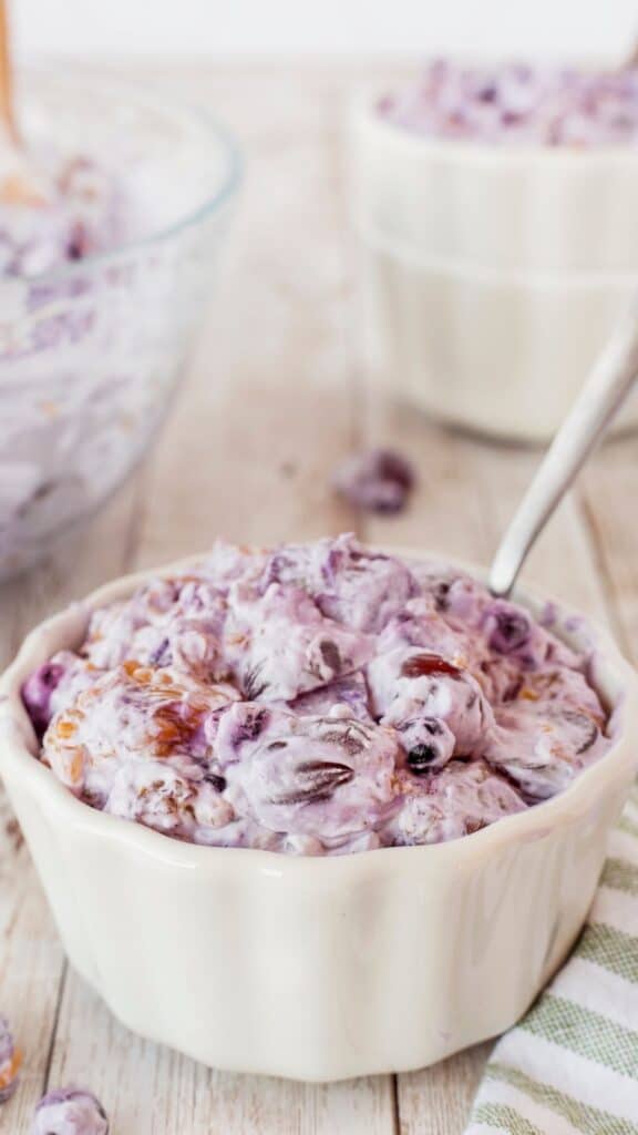 A creamy fruit salad with a purple hue is served in a white bowl on a wooden table. The salad contains visible grapes and nuts. A metal spoon rests inside the bowl. A partially visible bowl with more salad is in the background.