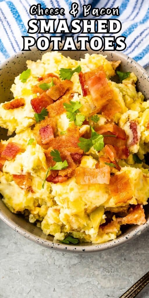 A bowl of easy smashed potatoes topped with crispy bacon bits and garnished with fresh herbs. The dish is labeled Cheese & Bacon Smashed Potatoes at the top of the image, with a striped towel visible in the background.