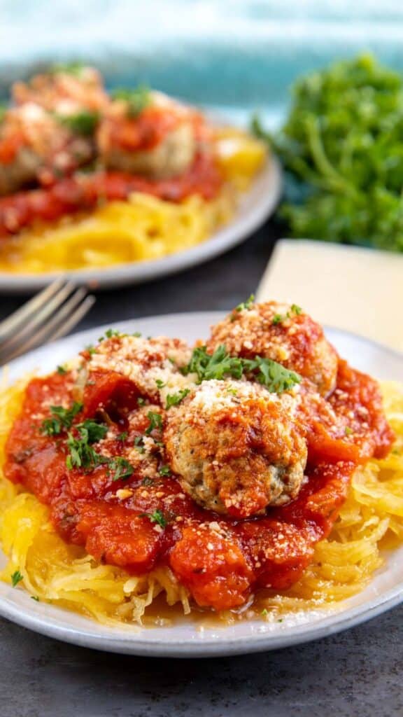 A plate of spaghetti squash topped with tomato sauce and meatballs, garnished with grated cheese and fresh parsley. Another plate with the same dish is slightly blurred in the background, with a sprig of fresh parsley nearby.