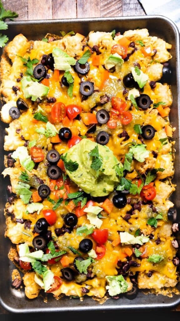 A baking tray filled with loaded nachos topped with melted cheese, black olives, cherry tomatoes, and avocado chunks. Garnished with cilantro and a dollop of guacamole in the center.