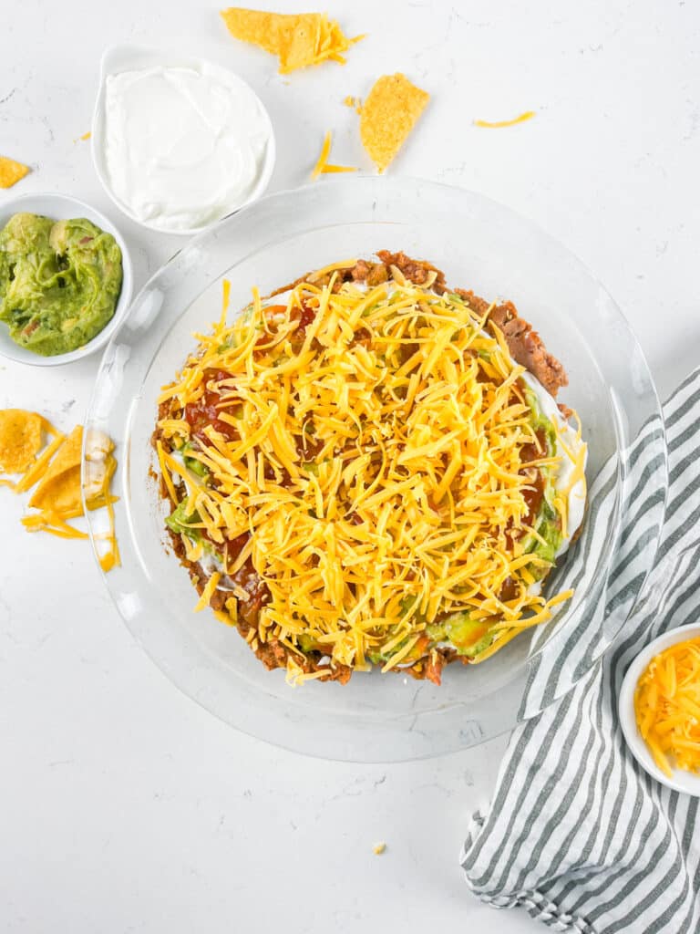 A plate of layered taco dip topped with shredded cheese, surrounded by small bowls of guacamole, sour cream, and extra cheese. Tortilla chips are scattered around, and a striped cloth napkin is nearby.