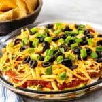 Glass dish with layered dip, including guacamole, salsa, shredded cheese, black olives, and chopped green onions. A bowl of tortilla chips is in the background. A striped cloth napkin is nearby.