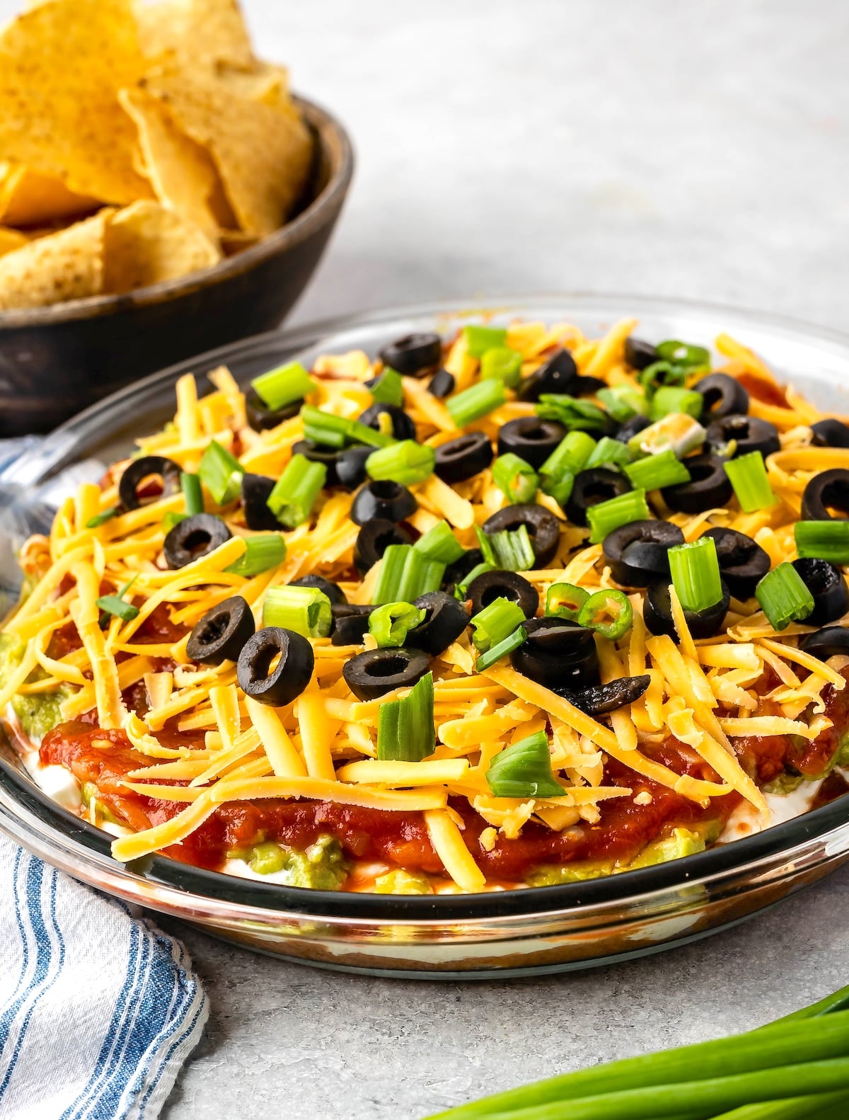 Glass dish with layered dip, including guacamole, salsa, shredded cheese, black olives, and chopped green onions. A bowl of tortilla chips is in the background. A striped cloth napkin is nearby.