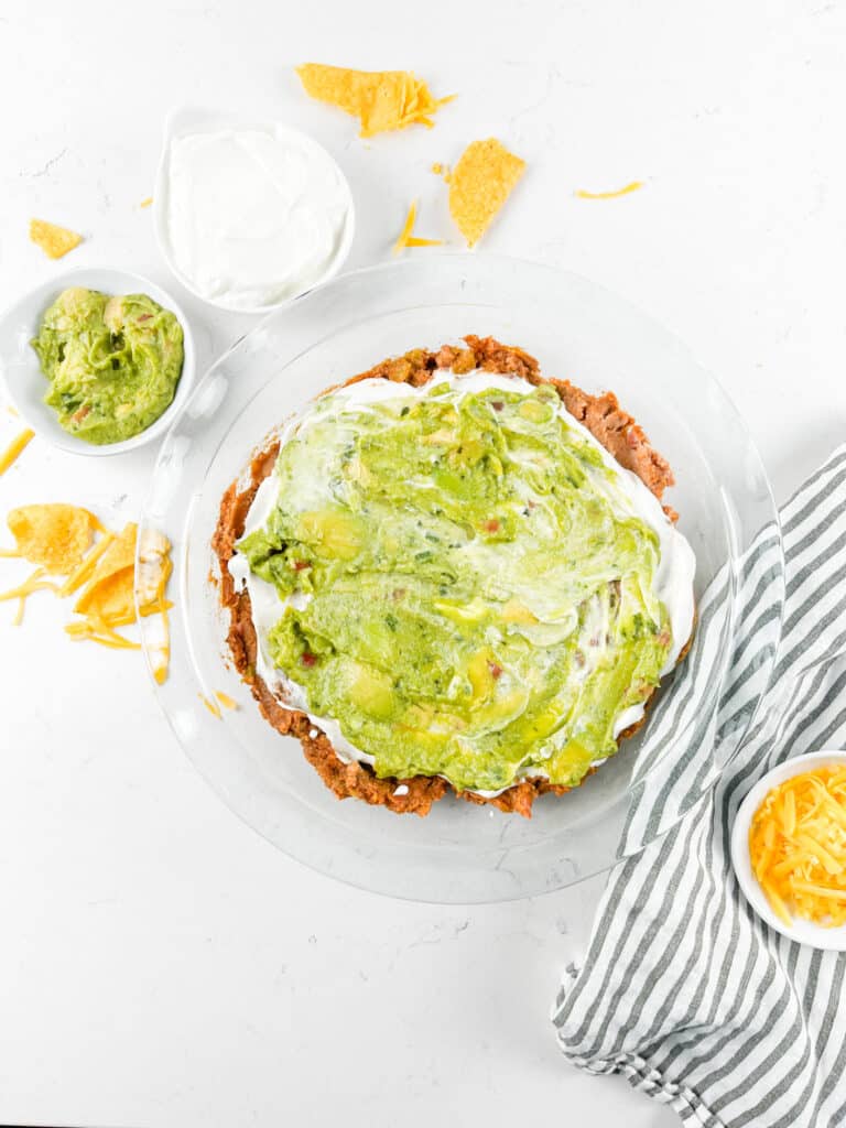 A layered dip in a glass dish with refried beans, sour cream, and guacamole on a white surface. Surrounding are small bowls of sour cream, guacamole, and shredded cheese. Tortilla chips are scattered nearby, with a striped cloth beside them.