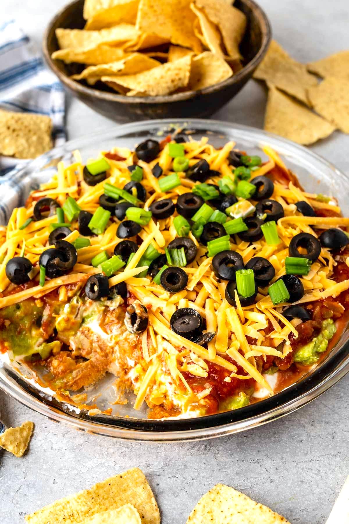 A glass dish filled with layered taco dip topped with shredded cheese, sliced black olives, and chopped green onions. Surrounding the dish are tortilla chips, some in a bowl and others scattered on a gray surface.