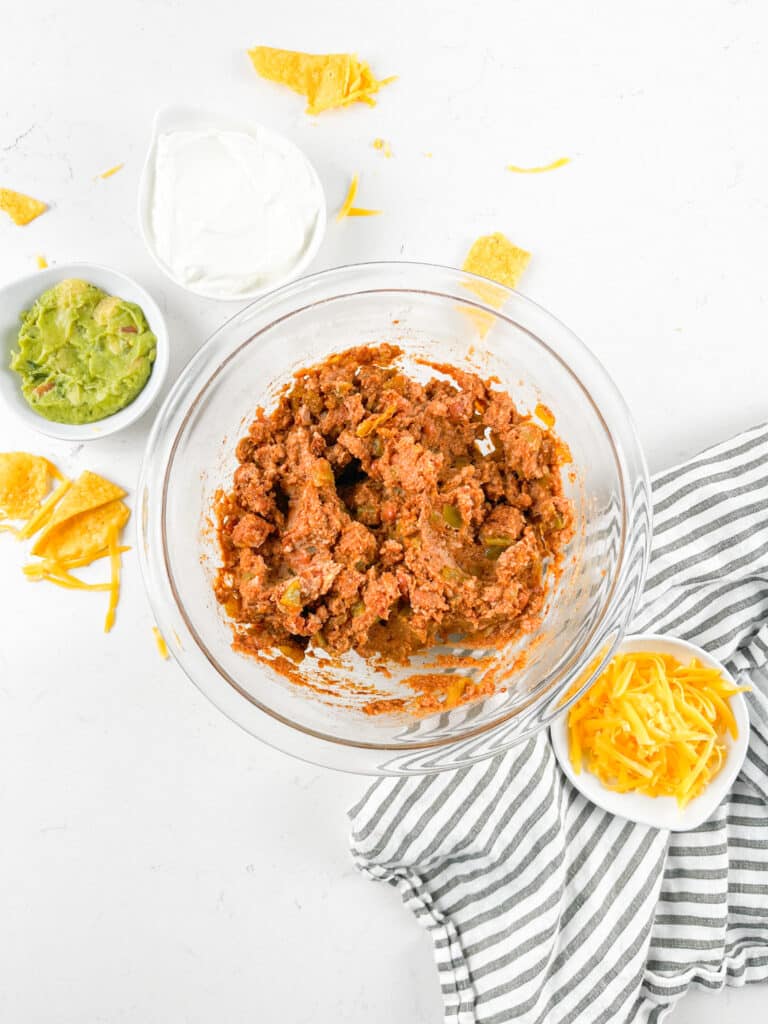 A glass bowl filled with a ground meat mixture is surrounded by small bowls of guacamole, sour cream, and shredded cheese. Tortilla chips are scattered around, with a striped cloth napkin nearby on a white surface.