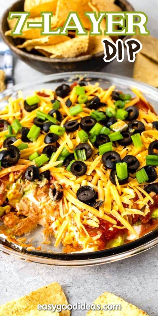 A plate of 7-layer dip topped with shredded cheese, sliced black olives, and chopped green onions. In the background, there are tortilla chips and a text overlay reads 7-Layer Dip.