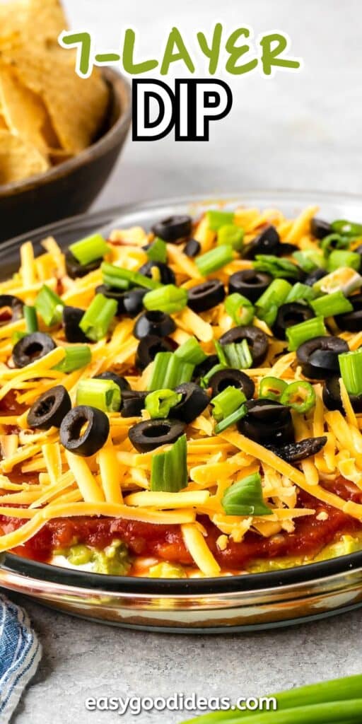 A plate filled with a seven-layer dip topped with sliced black olives, shredded cheese, and chopped green onions. A bowl of tortilla chips is in the background. The text 7-Layer Dip is at the top, and easygoodideas.com is at the bottom.