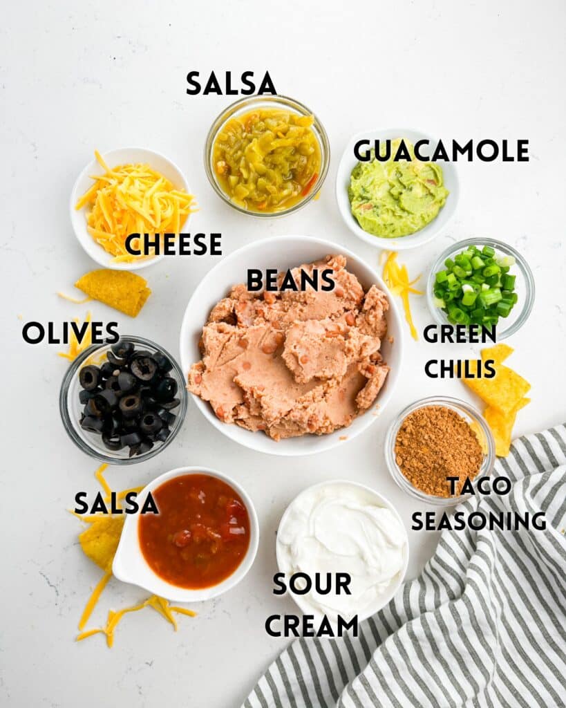 A variety of taco dip ingredients are arranged in bowls on a table: beans, salsa, guacamole, shredded cheese, green chilies, olives, sour cream, and taco seasoning. A striped napkin and tortilla chips are nearby.