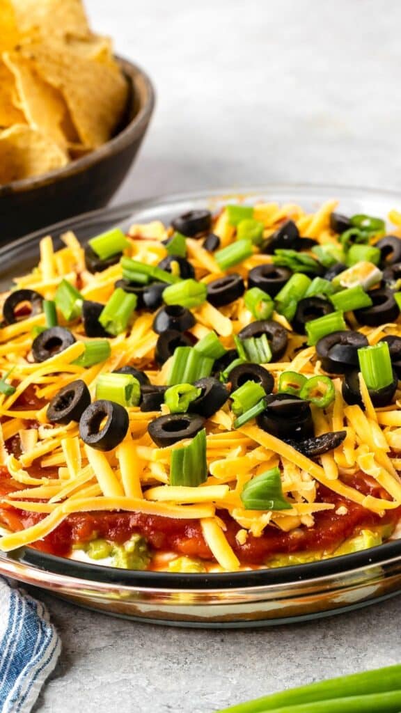 A glass dish filled with a layered dip topped with shredded cheese, sliced black olives, and chopped green onions. A bowl of tortilla chips is partially visible in the background.