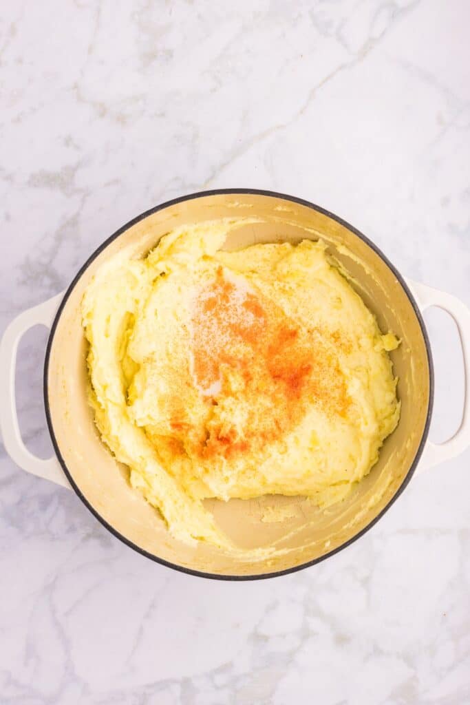 A pot filled with creamy mashed potatoes, topped with a sprinkle of orange spices, likely paprika. The pot has white handles and is placed on a marble surface.