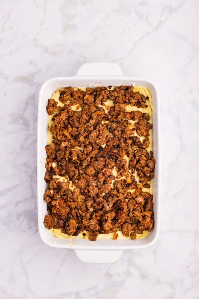 White rectangular baking dish on a marble surface, filled with a layered casserole topped with crumbled cooked ground meat, likely beef or sausage, and a visible creamy layer beneath.