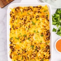 A casserole dish filled with a cheesy, baked ground beef and onion dish, garnished with parsley. Nearby are grated cheese, an onion, salt and pepper shakers, fresh parsley, and a small bowl of seasoning on a marble countertop.