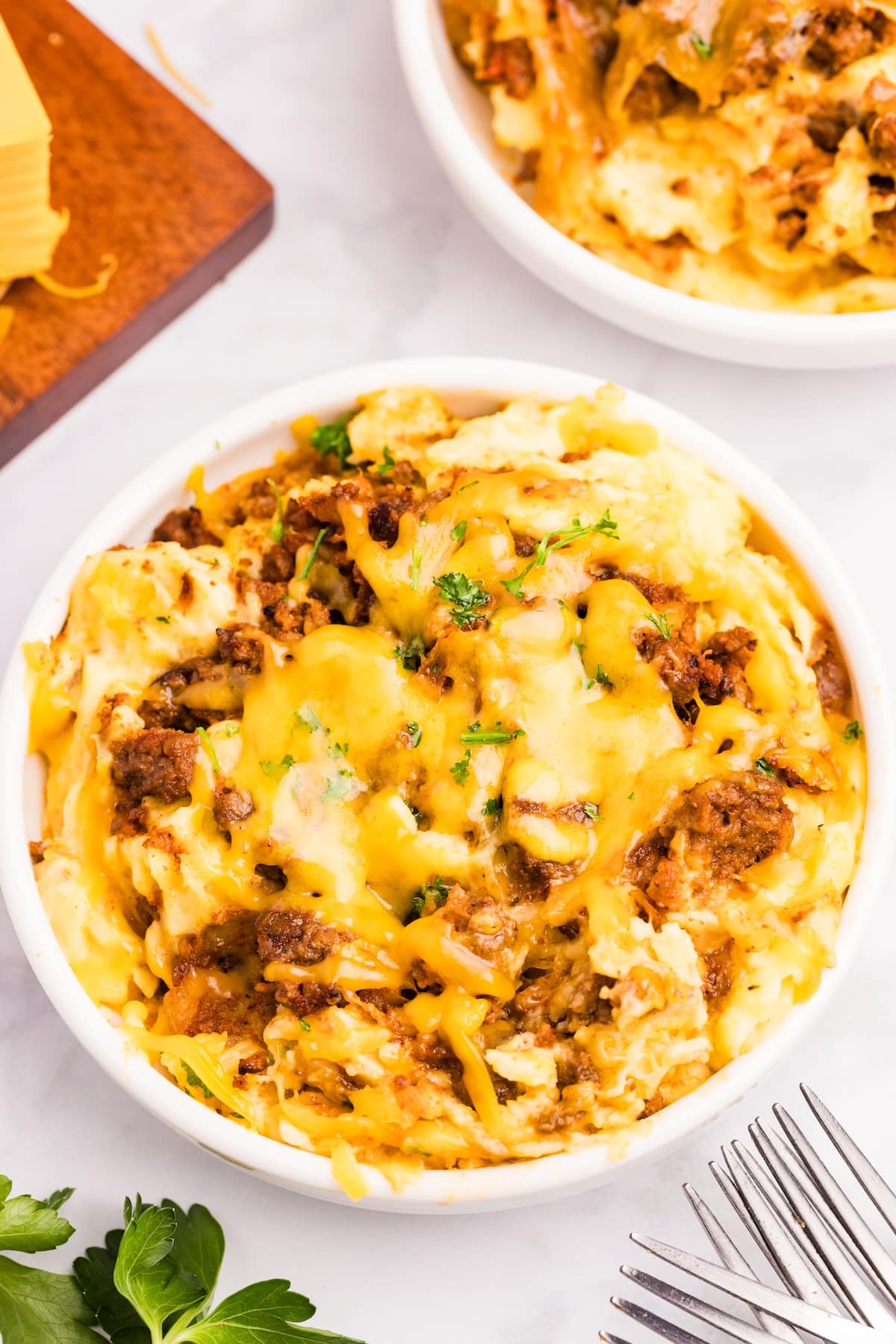 A bowl of cheesy ground beef and potato casserole topped with melted cheese and chopped parsley. There are forks and parsley sprigs nearby, with a partial view of another bowl in the background.