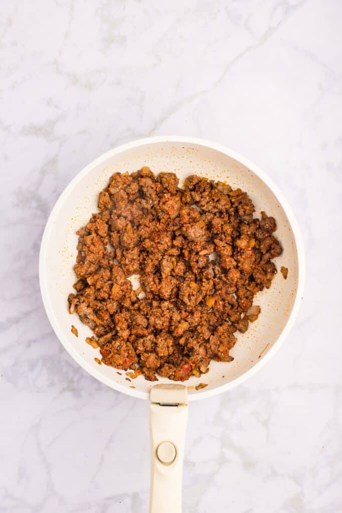 A white skillet on a marble surface contains cooked, crumbled seasoned beef with visible spices, creating a rich, brown texture.