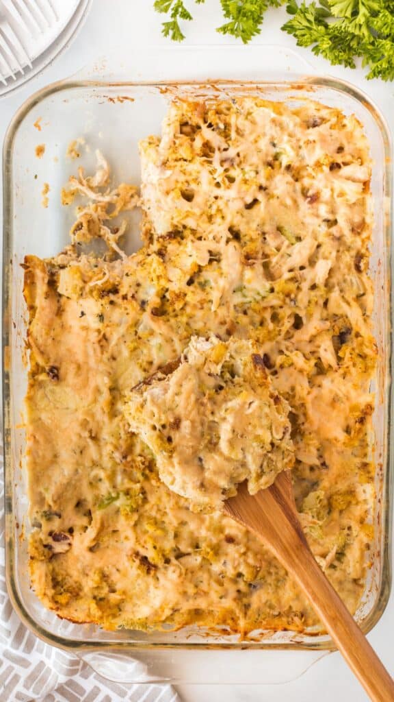 A rectangular glass baking dish containing a partially served cheesy chicken and rice casserole, with a wooden spoon scooping a portion. The top is golden and slightly crispy, with a sprinkle of herbs. Fresh parsley is visible in the background.