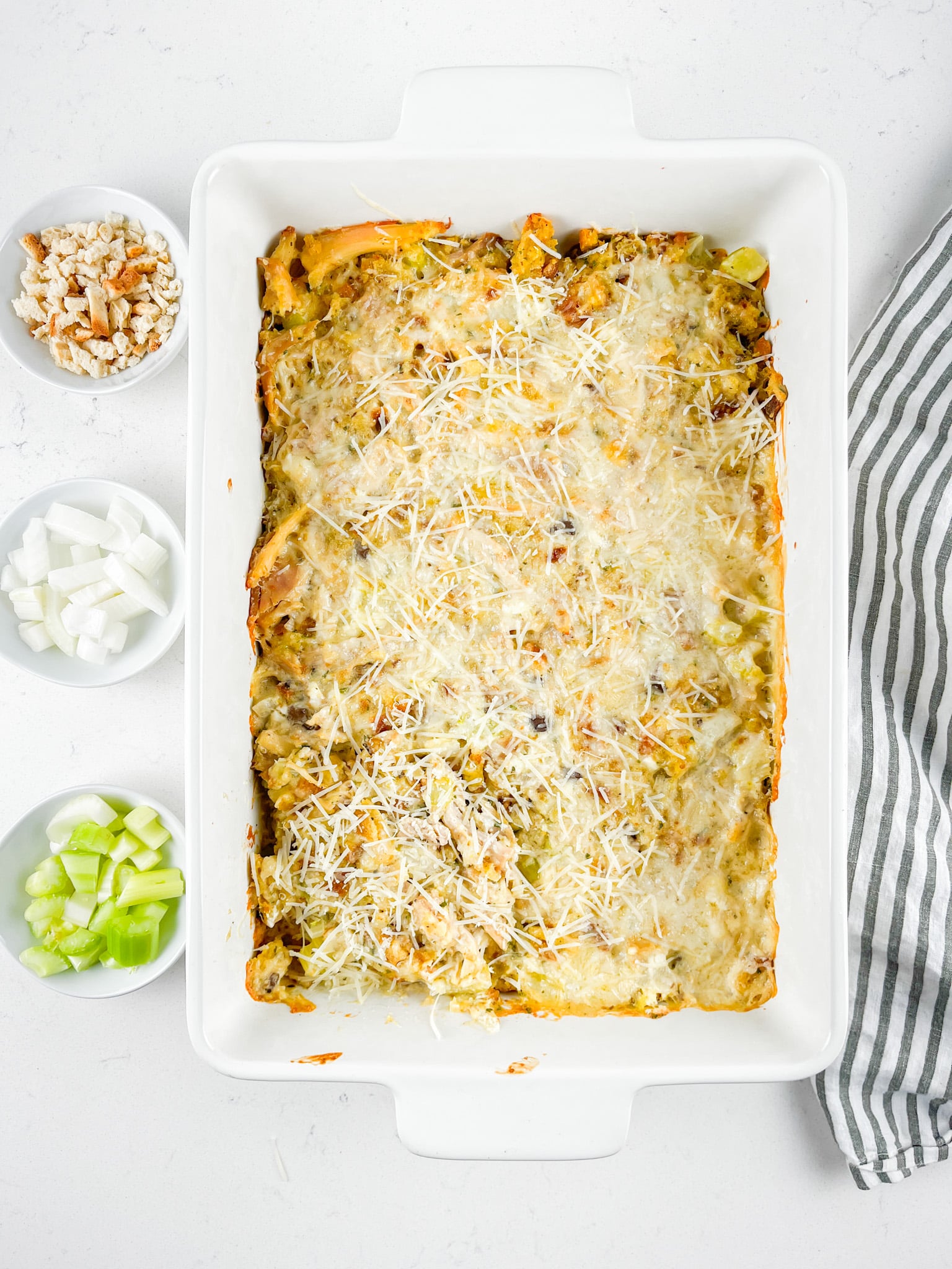 A baked casserole topped with grated cheese in a white dish. To the side, there are small bowls containing chopped celery, onions, and bread cubes. A striped cloth napkin is partially visible on the right.