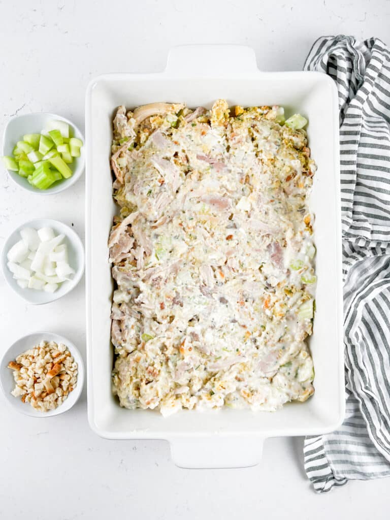 A rectangular baking dish filled with a creamy chicken casserole mixture. Ingredients including chopped celery, onions, and breadcrumbs are placed in small bowls alongside. A striped kitchen towel is nearby on a light countertop.