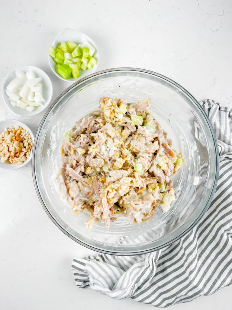 A glass bowl containing a mixture of shredded chicken, mayonnaise, celery, and walnuts. Next to the bowl are small dishes with cut celery, chopped onion, and additional walnuts on a white surface, accompanied by a striped cloth.