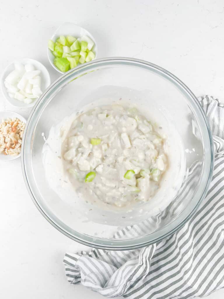 A glass bowl filled with a creamy white mixture containing chopped celery and onions. Small bowls with chopped celery, onions, and walnuts are nearby. A gray and white striped cloth is draped beside the bowl on a white surface.