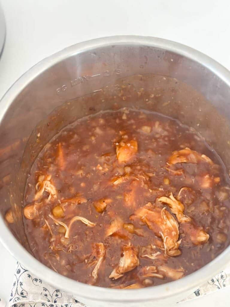 A pot filled with shredded chicken in a rich, dark brown sauce, possibly barbecue or stew. The pot is placed on a white surface with a patterned cloth underneath.