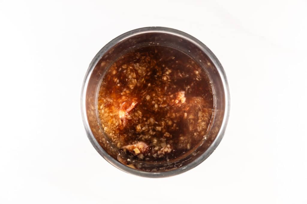 A glass jar filled with a reddish-brown liquid containing visible minced garlic and small pieces of onion, viewed from above on a white background.