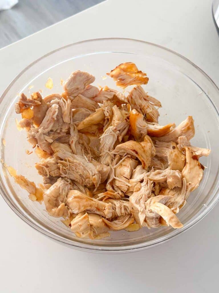 A clear glass bowl filled with shredded pieces of cooked chicken, lightly seasoned and showing some golden brown edges. The bowl is placed on a white surface, with part of a dark object visible in the background.