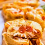 Close-up of sliced pizza bread with visible mozzarella cheese and small pepperoni slices on a wooden board. The bread is golden-brown and stuffed with melted cheese and pepperoni, creating a gooey texture. A whole loaf is blurred in the background.